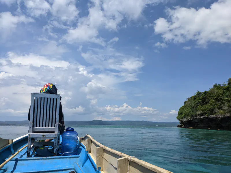 Person sitting on a chair at the front of a blue boat with clear ocean water and distant islands under a partly cloudy sky.
