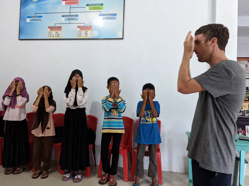 Man leading a group of children in an activity where they cover their faces with their hands indoors.