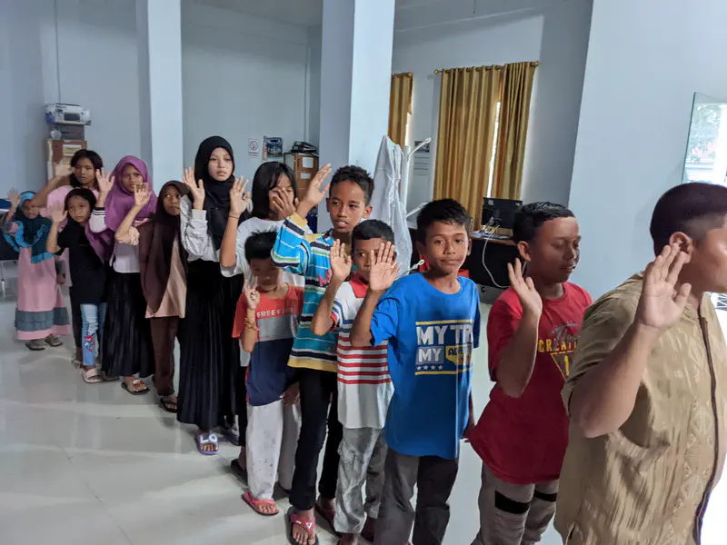 Group of children standing in a line indoors with one hand raised, participating in an activity.