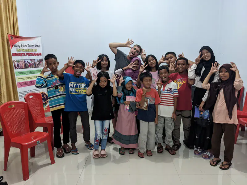 Group of children and an adult indoors smiling and making playful hand gestures in front of a poster and red chairs.