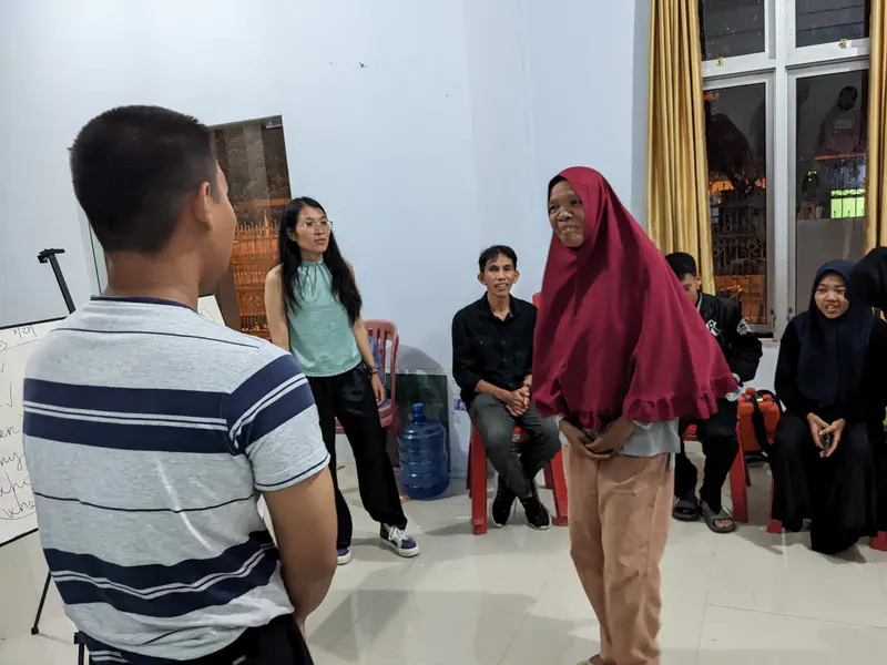 Girl in a red headscarf standing and smiling during an indoor activity while others watch and participate.