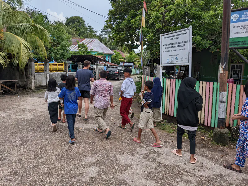 Group of children walking with an adult down a village street past houses and signboards.