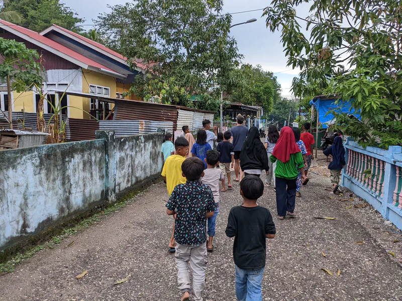 Group of children and adults walking together down a village street lined with houses and trees.