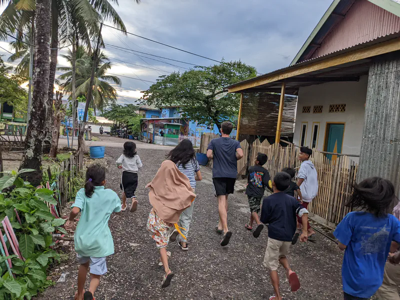 Group of children running with an adult down a village street lined with houses and palm trees toward the sunset.