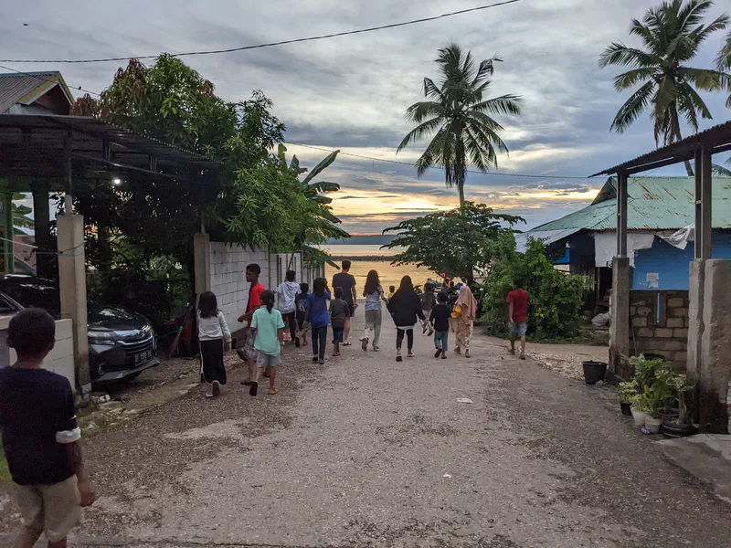 Group of children and adults walking down a village street toward the sea at sunset with palm trees around.