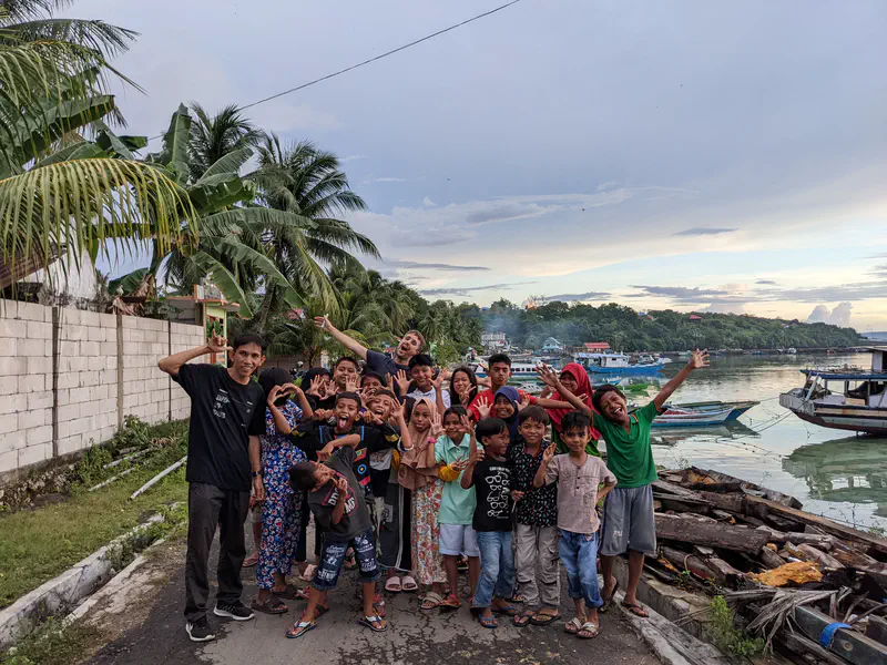 Group of children and adults posing playfully near the waterfront with palm trees and boats in the background.