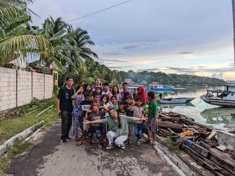 Group of children and adults gathered near the waterfront with boats and palm trees in the background, smiling and posing.