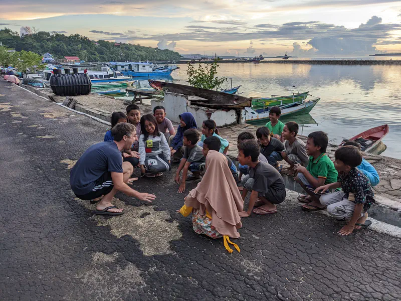 Man crouching and talking with a group of children sitting together by the waterfront with boats and a sunset in the background.