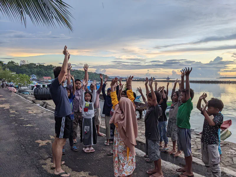 Man leading children in raising their arms during an outdoor activity by the waterfront at sunset.