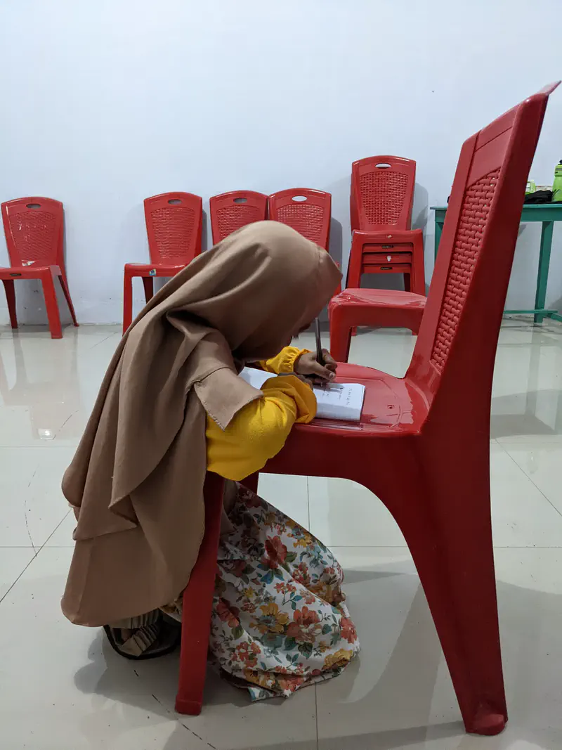 Girl in a headscarf kneeling on the floor and writing on paper placed on a red plastic chair indoors.