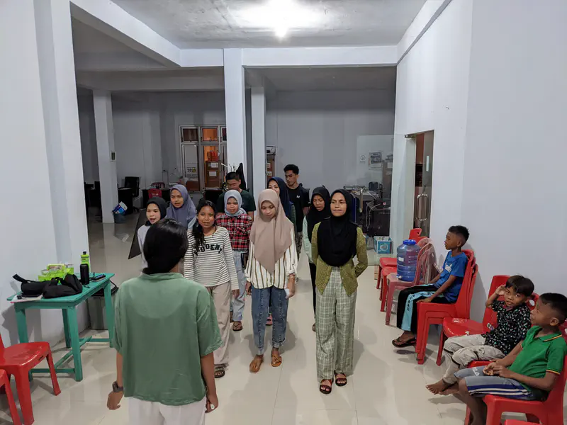Teenagers standing in rows indoors facing a person leading an activity, while younger children sit and watch.