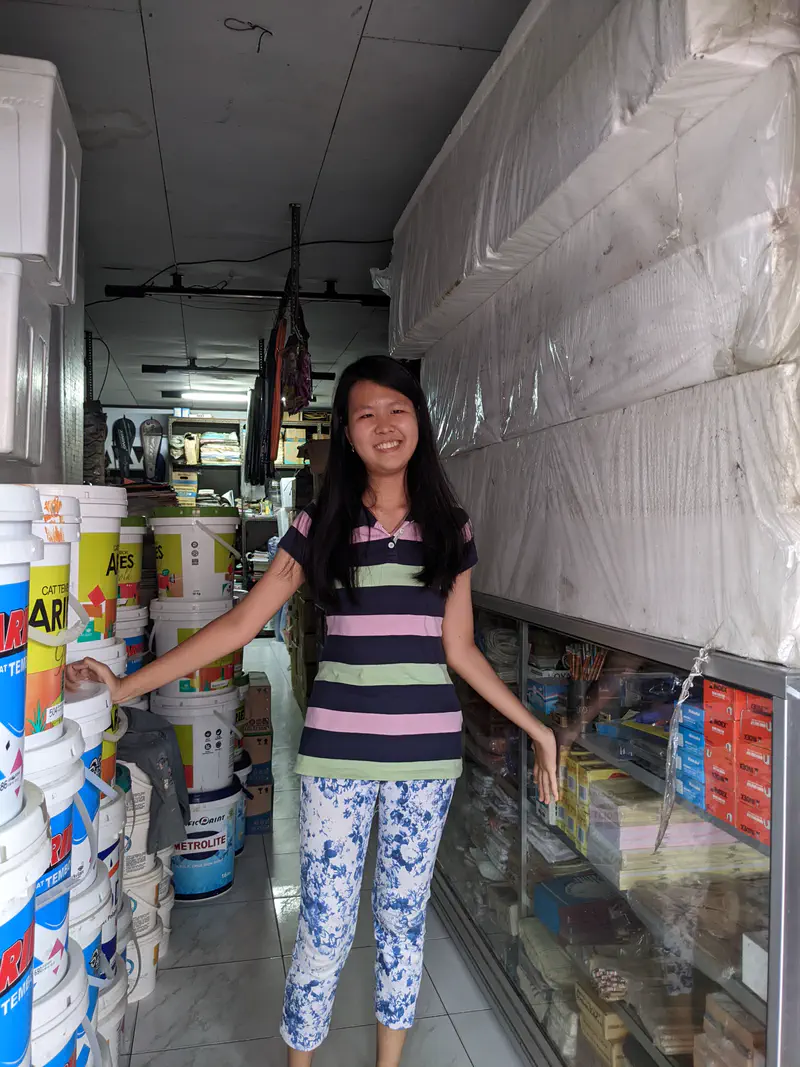 Smiling woman standing inside a shop with paint buckets stacked on one side and mattresses wrapped in plastic on the other.