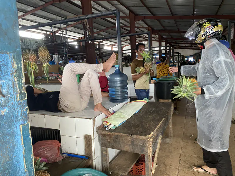 Man selling pineapples to a customer wearing a helmet and raincoat inside a market.