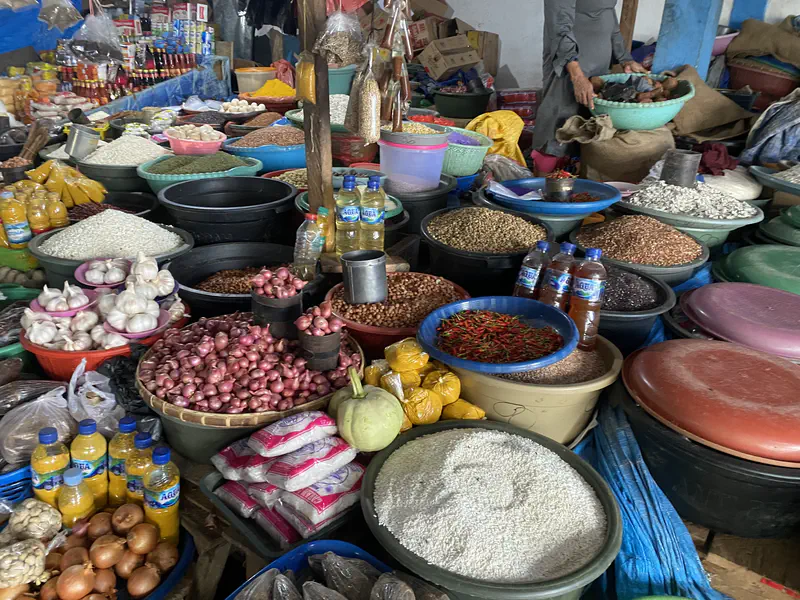 Market stall with baskets of onions, garlic, chilies, grains, and bottled cooking oil.