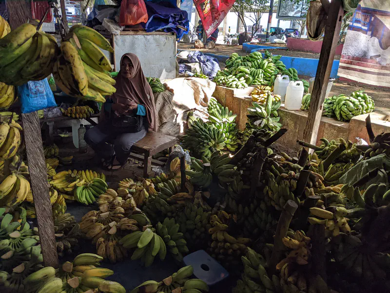 Woman sitting among piles of bananas at a shaded market stall.