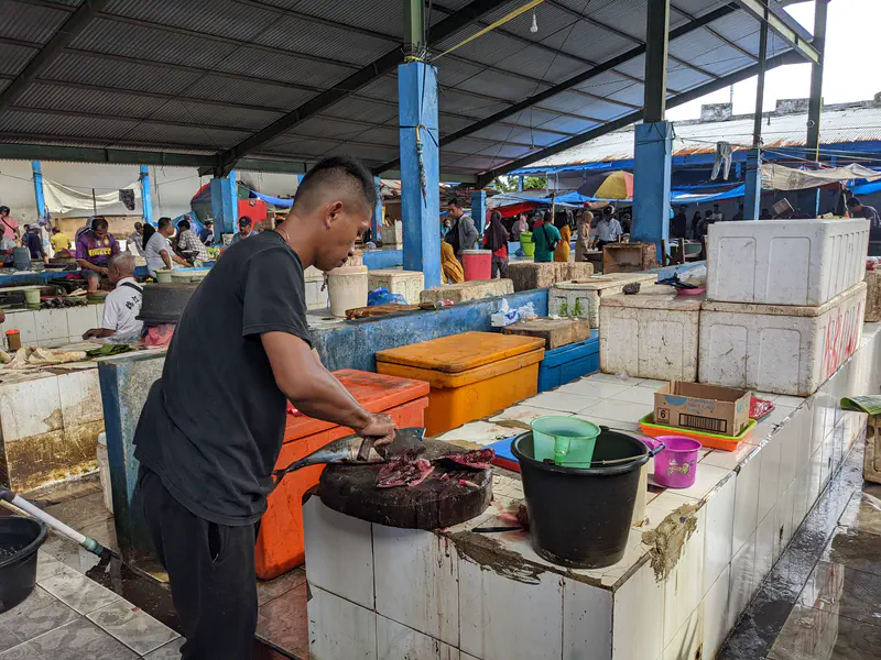 Man chopping fish on a wooden block inside a busy covered market.
