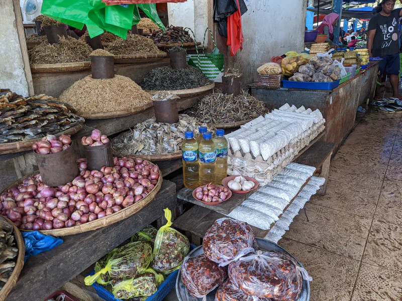 Market stall with onions, garlic, dried fish, tempeh, and bottled cooking oil on display.