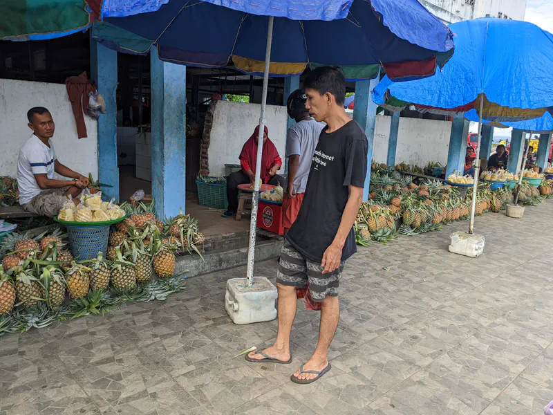 Man walking past pineapple vendors under blue umbrellas at a market.