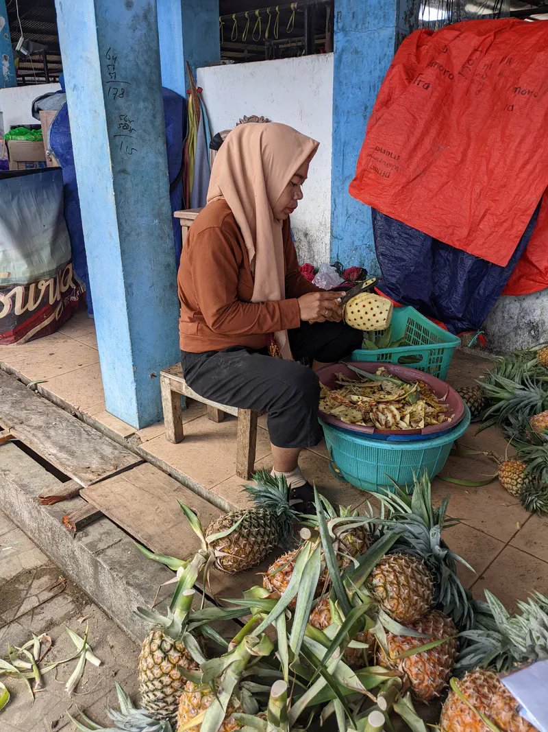 Woman peeling a pineapple while sitting beside baskets of fruit at a market stall.