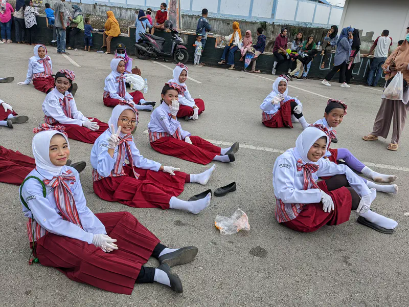 Group of girls in red and white uniforms sitting on the road smiling during a parade break.