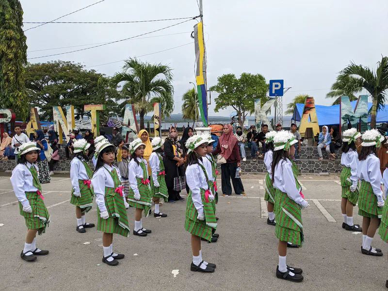 Girls in green plaid skirts and white blouses with flower hats marching in a parade.