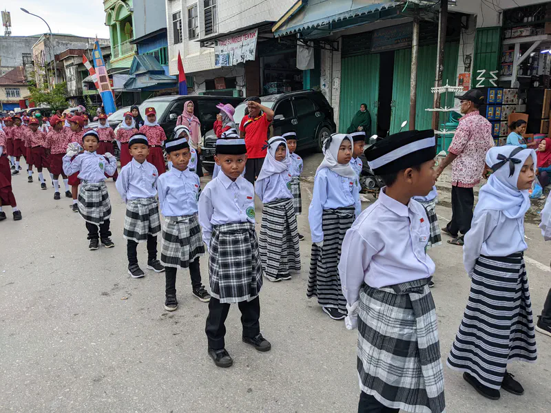 Boys and girls in white shirts and black plaid sarongs marching in a parade.