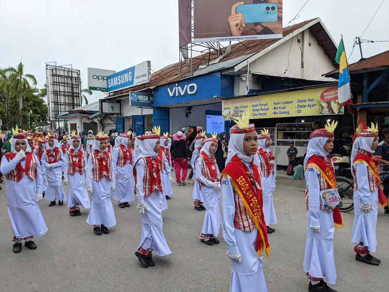 Girls in white uniforms with red plaid vests and sashes marching in a parade.