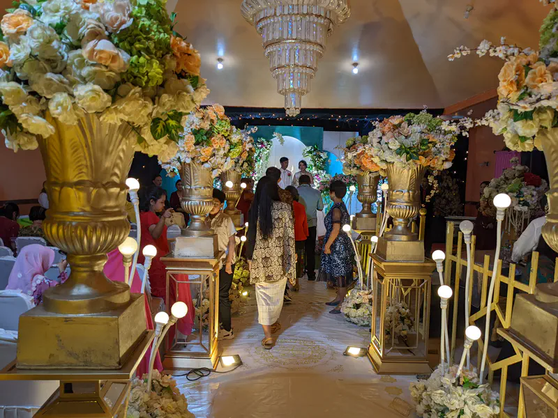 Wedding guests walking down a decorated aisle with flower arrangements and chandeliers toward the bride and groom on stage.