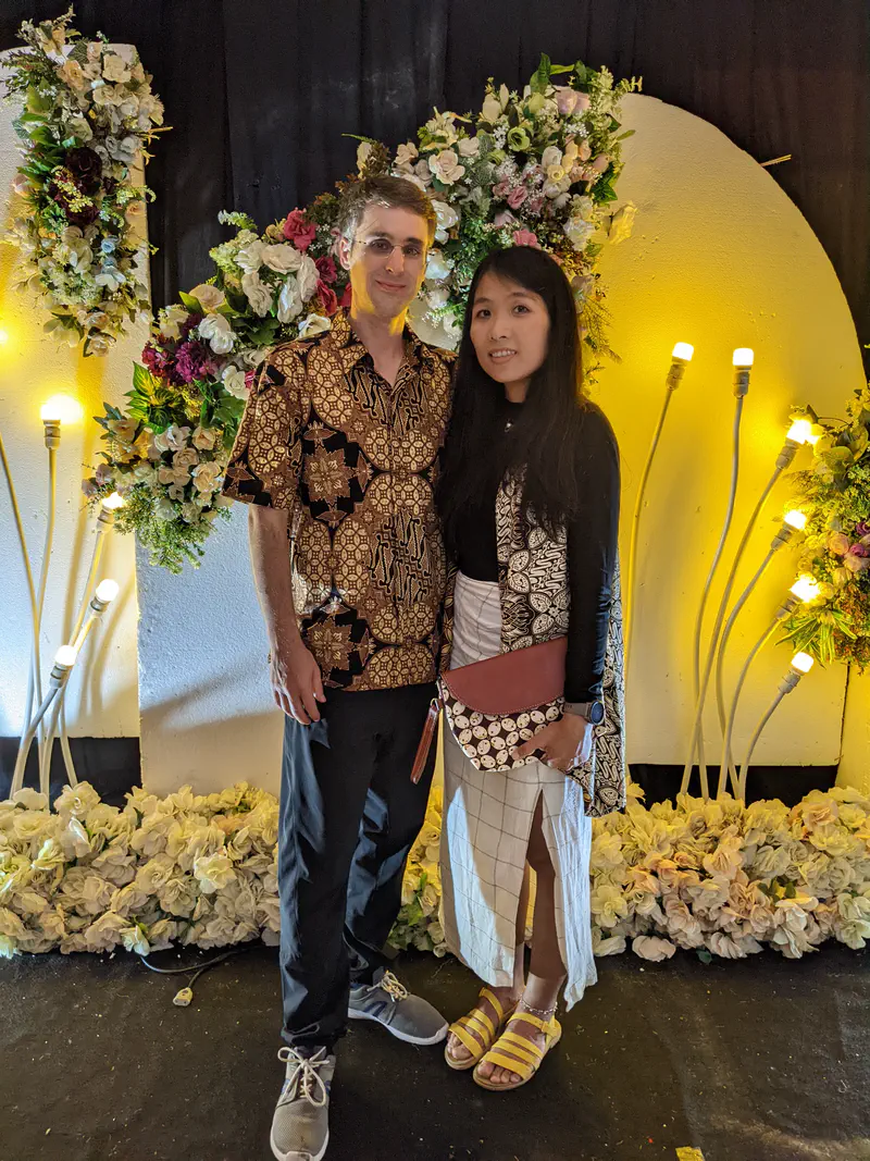 Couple dressed in batik and formal attire posing in front of a wedding floral backdrop.