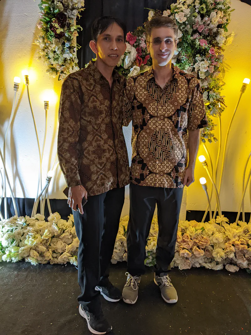 Two men wearing batik shirts posing together in front of a wedding floral backdrop.