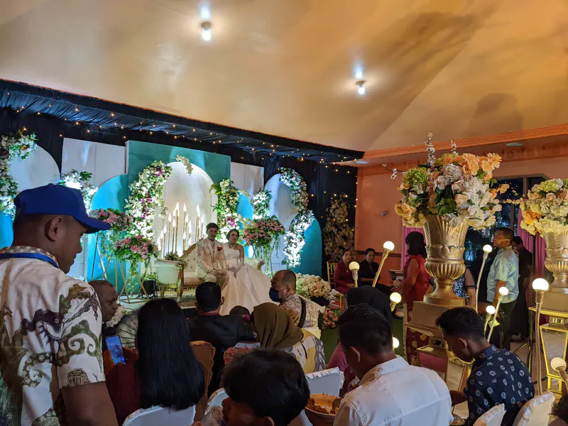 Bride and groom sitting on a decorated stage at their wedding while guests are seated and dining.