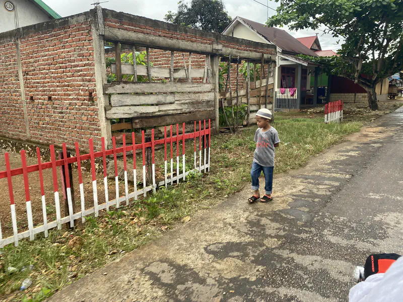 A young boy wearing a cap, grey t-shirt, and sandals walks along a village road beside a red and white fence and brick houses.