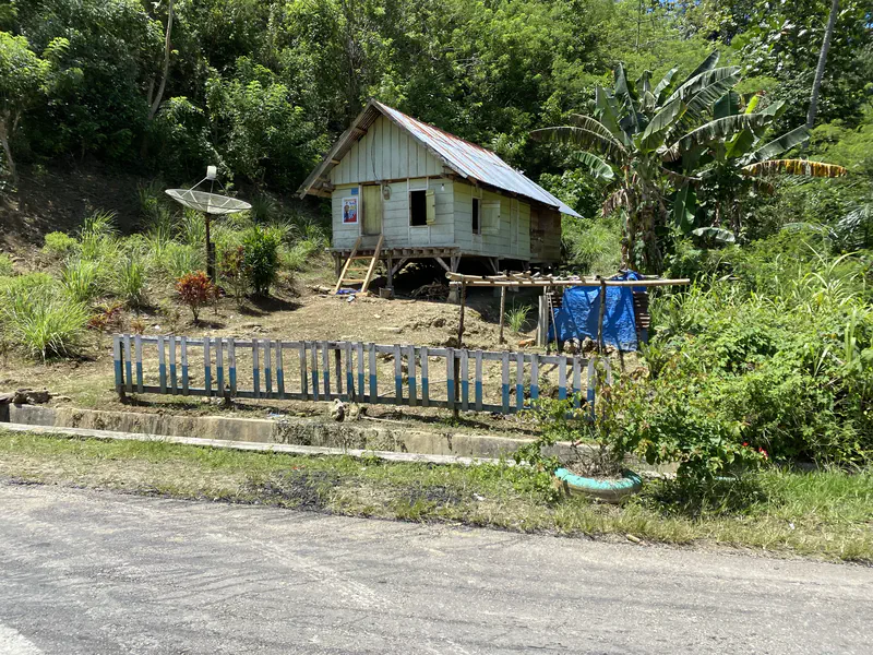 Small stilt house with a tin roof and satellite dish on a hillside surrounded by greenery.