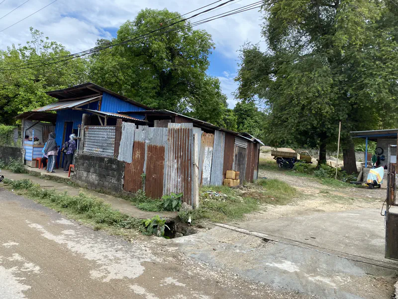 People standing outside a blue roadside shack with corrugated metal walls near a tree and dirt path.
