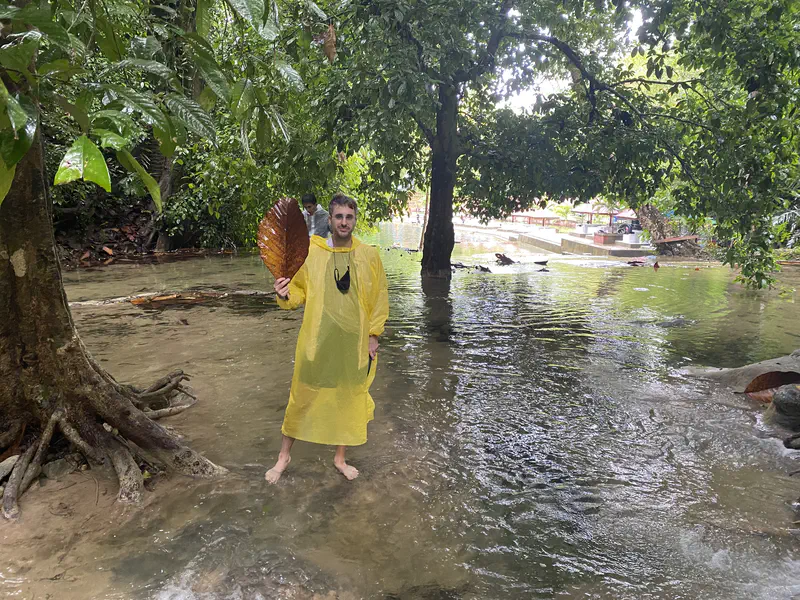 Man in a yellow rain poncho holding a large brown leaf while standing barefoot in shallow water among trees.