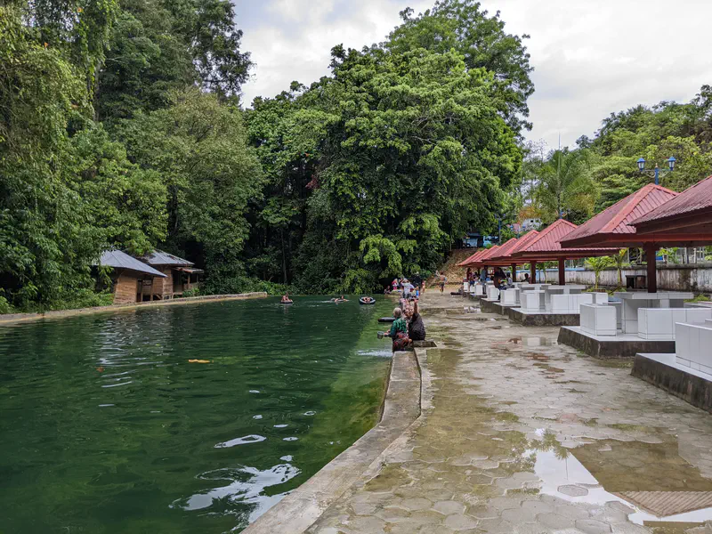 People sitting and swimming by a large outdoor pool surrounded by trees and red-roofed shelters.
