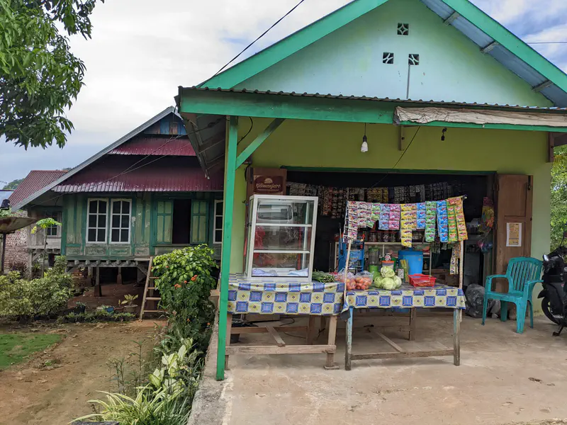 Small village shop with vegetables and packaged goods displayed in front of a green house.