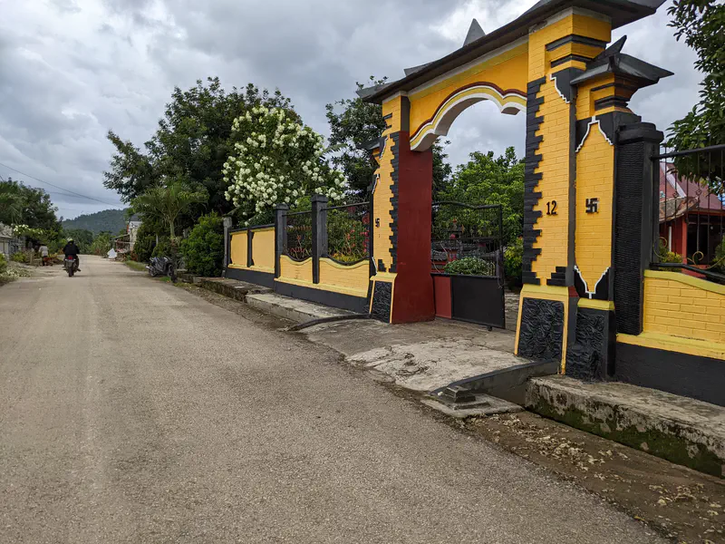 Motorcyclist on a village road passing a large yellow and black gate with decorative pillars.