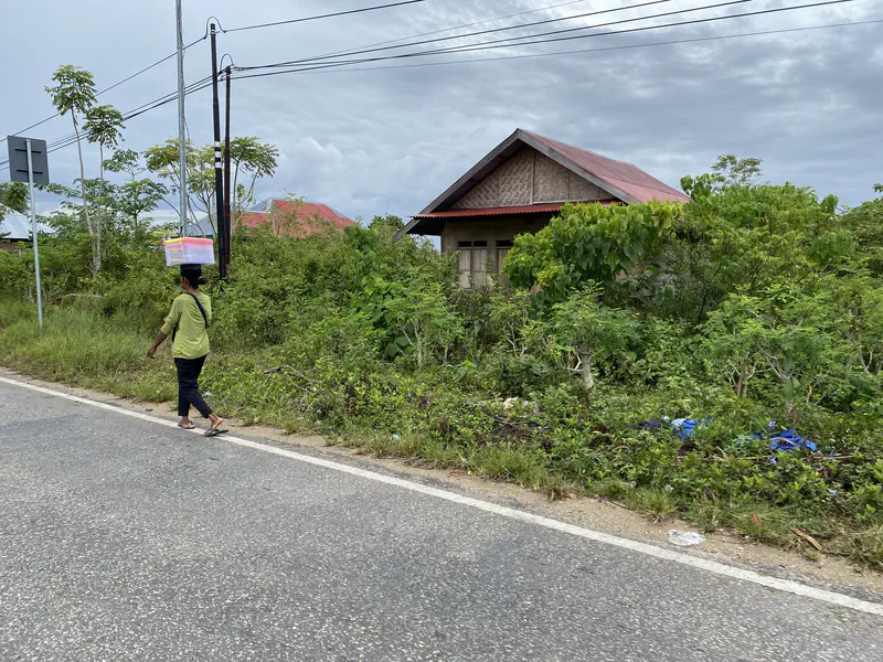 Person walking barefoot on a roadside carrying a plastic container balanced on their head, with a house and greenery in the background.