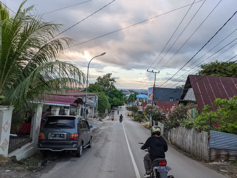 Motorcyclists riding down a village street lined with houses and trees at sunset.