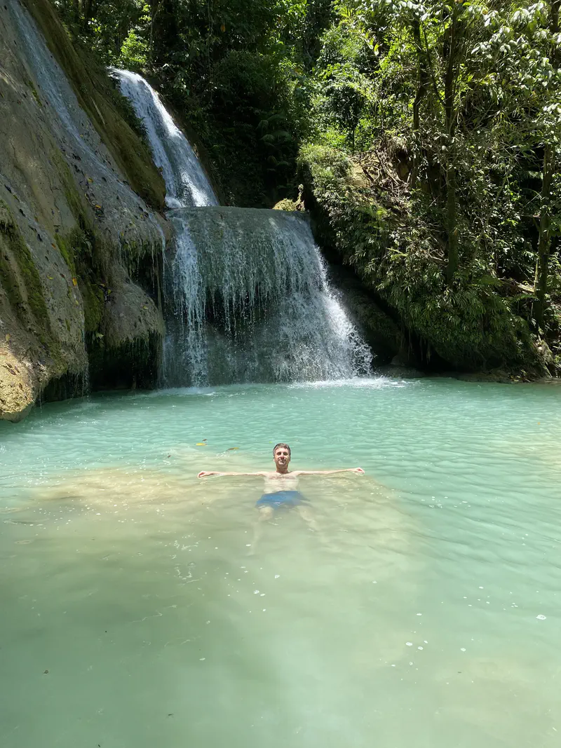 Person floating on their back with arms wide in turquoise water at the base of a jungle waterfall.