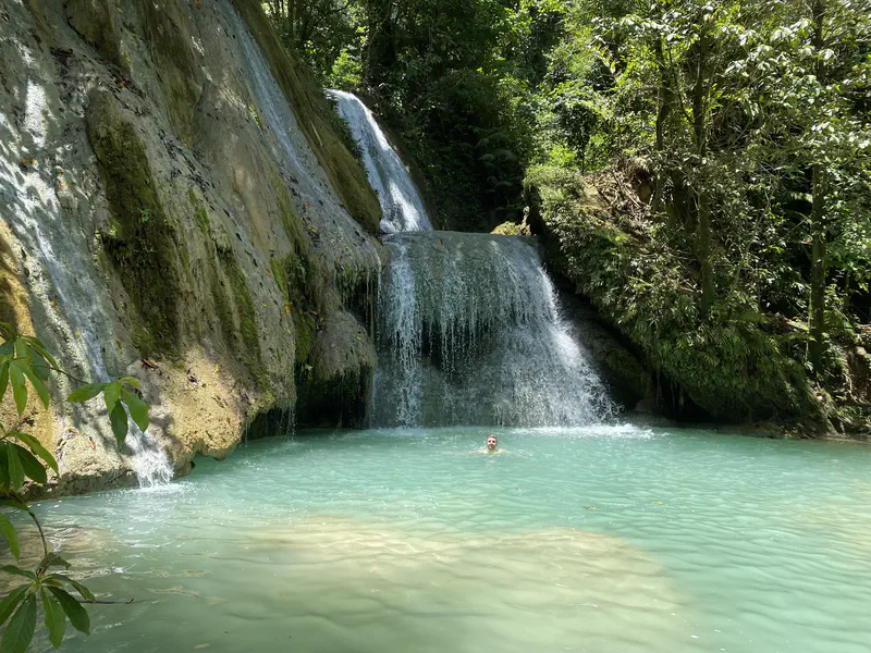 Person swimming in a turquoise pool beneath a cascading jungle waterfall with moss-covered rocks.