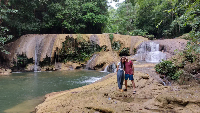 Couple standing on rocks in front of a multi-tiered waterfall surrounded by forest.