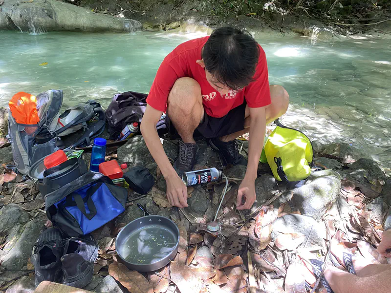 Person in a red shirt preparing a meal with camping gear beside a turquoise river.