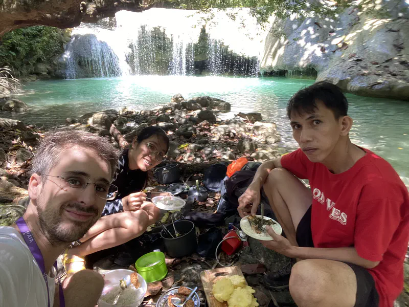 Three people eating a picnic meal beside a turquoise waterfall pool.
