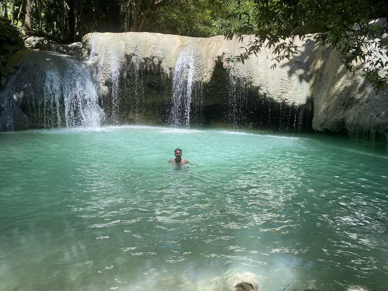 Person swimming in a turquoise pool beneath a small waterfall surrounded by rocks and trees.