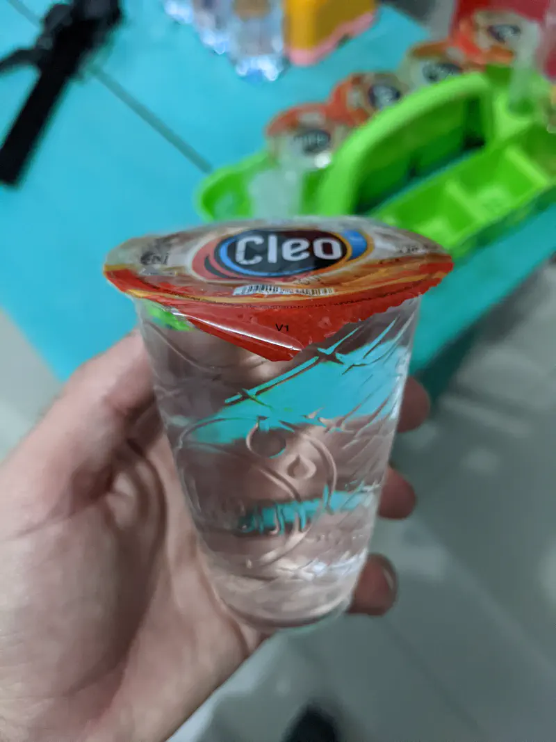 Hand holding a sealed plastic cup of Cleo brand drinking water.