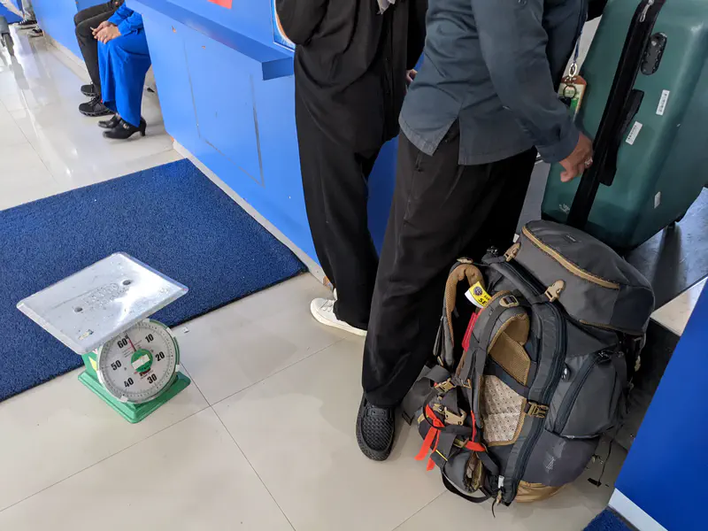 Travelers with backpacks and suitcases standing near a counter with a luggage scale on the floor.