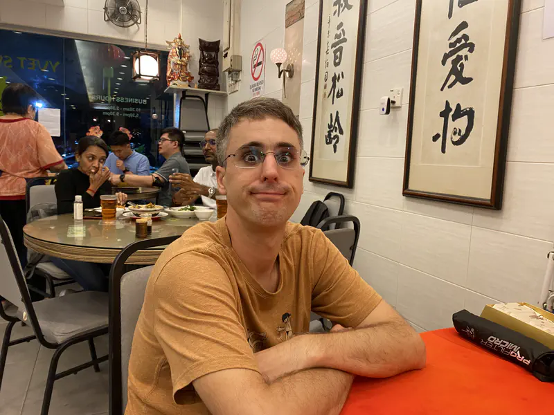 Person sitting at a round table in a Chinese restaurant, making a funny face, with other diners and calligraphy scrolls on the walls in the background.