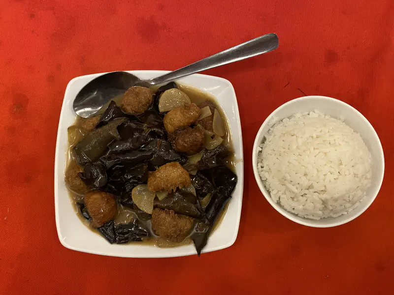 Close-up of a meal with a bowl of white rice and a plate of stir-fried vegetables and tofu in brown sauce on a red tablecloth.